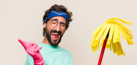 Un homme avec un bandeau qui tiens un balai espagnol et qui porte des gants de nettoyage roses ainsi qu'un tee-shirt bleu pastel